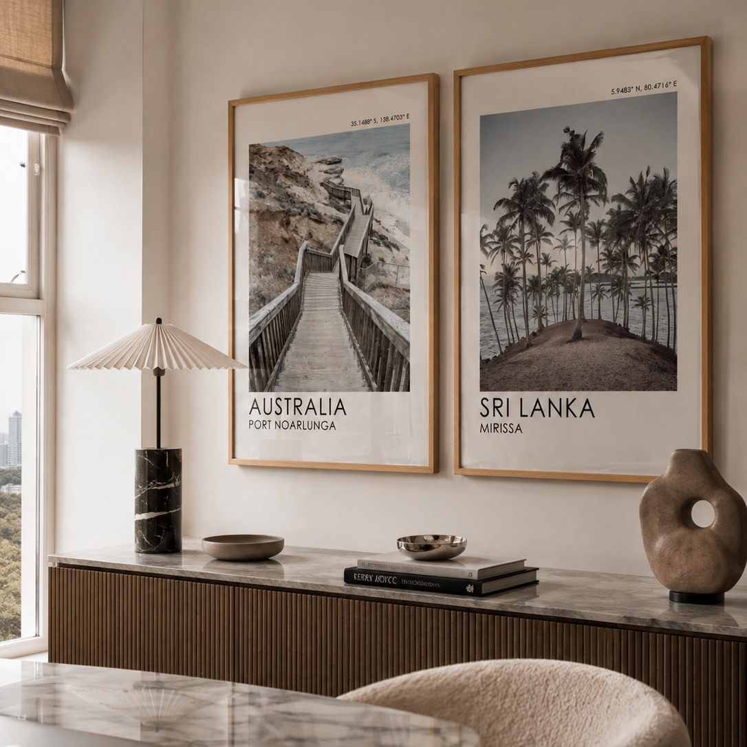 Two framed travel posters hanging on a beige wall above a marble console table in a modern interior. The left poster reads 'AUSTRALIA' and shows a bridge over water; the right poster reads 'SRI LANKA' and shows palm trees and a horse-drawn carriage.