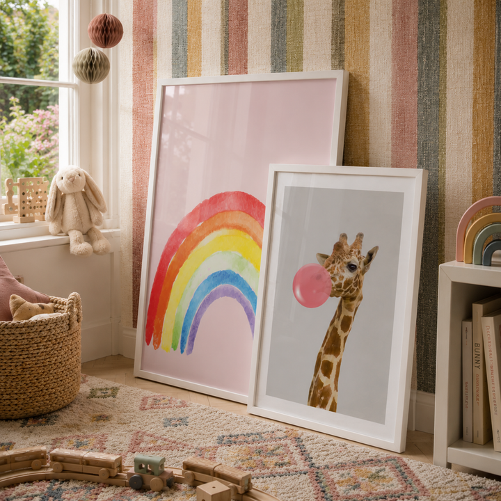 Two framed children's art prints leaning against a striped wall in a styled nursery. The larger print features a colorful rainbow on a soft pink background, and the smaller print shows a giraffe blowing a pink bubblegum bubble.