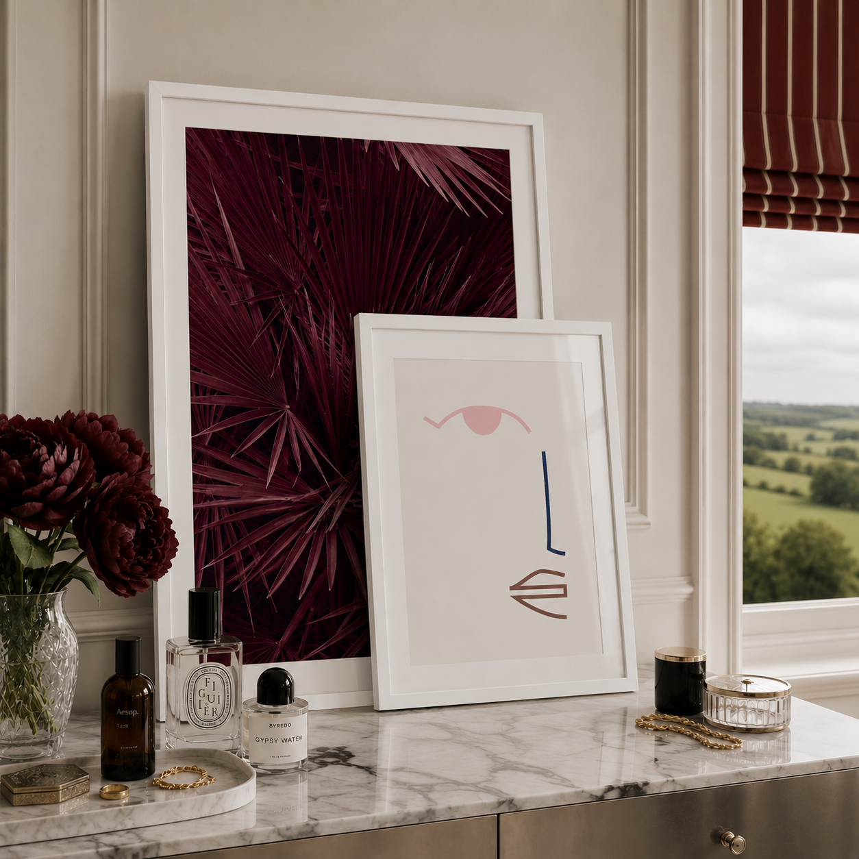 Two framed abstract botanical art prints displayed on a marble vanity table in a styled room. One print features deep burgundy palm leaves, and the other is a minimalist abstract design with pink, blue, and brown shapes on a white background.