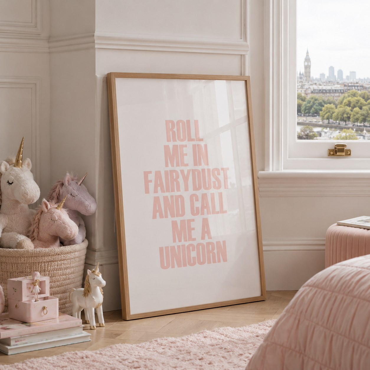 A framed typographic wall art print leaning against a cream wall in a styled bedroom. The print has pink text reading 'ROLL ME IN FAIRYDUST AND CALL ME A UNICORN' on a white background with a light wood frame.