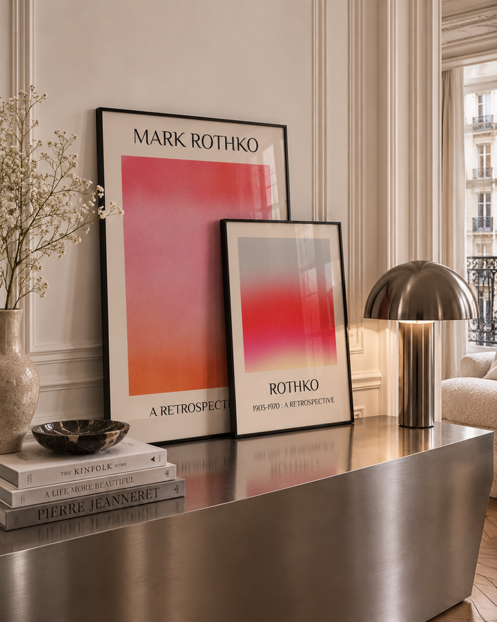 Two framed Mark Rothko art prints with abstract pink, red, and orange color fields are displayed on a metallic console table in a styled interior setting with a lamp, books, and a vase.