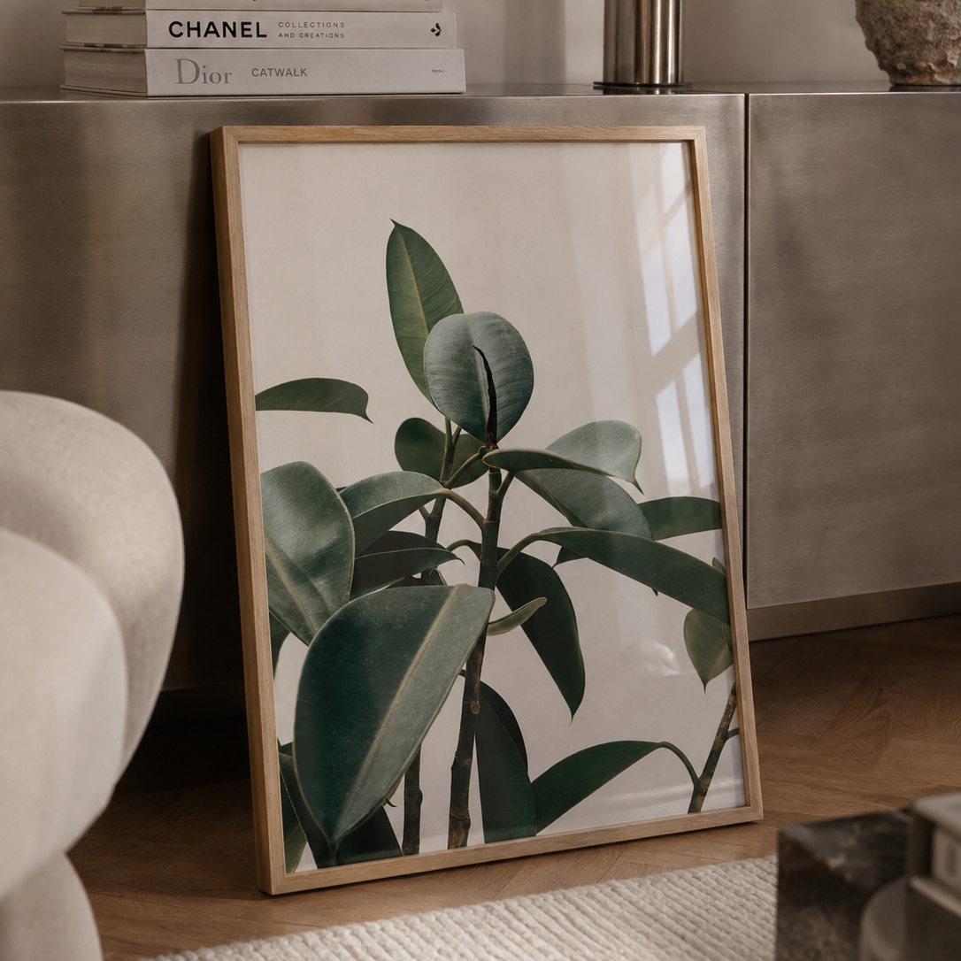 A framed botanical art print of a green rubber plant in a light wood frame, leaning against a metallic sideboard in a modern, neutral-toned room.