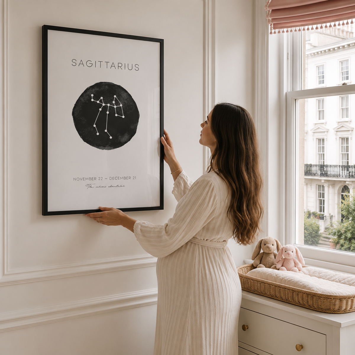 A woman in a cream ribbed dress hangs a framed Sagittarius constellation art print on a white wall next to a window. The print features a black circle with white constellation lines and text.