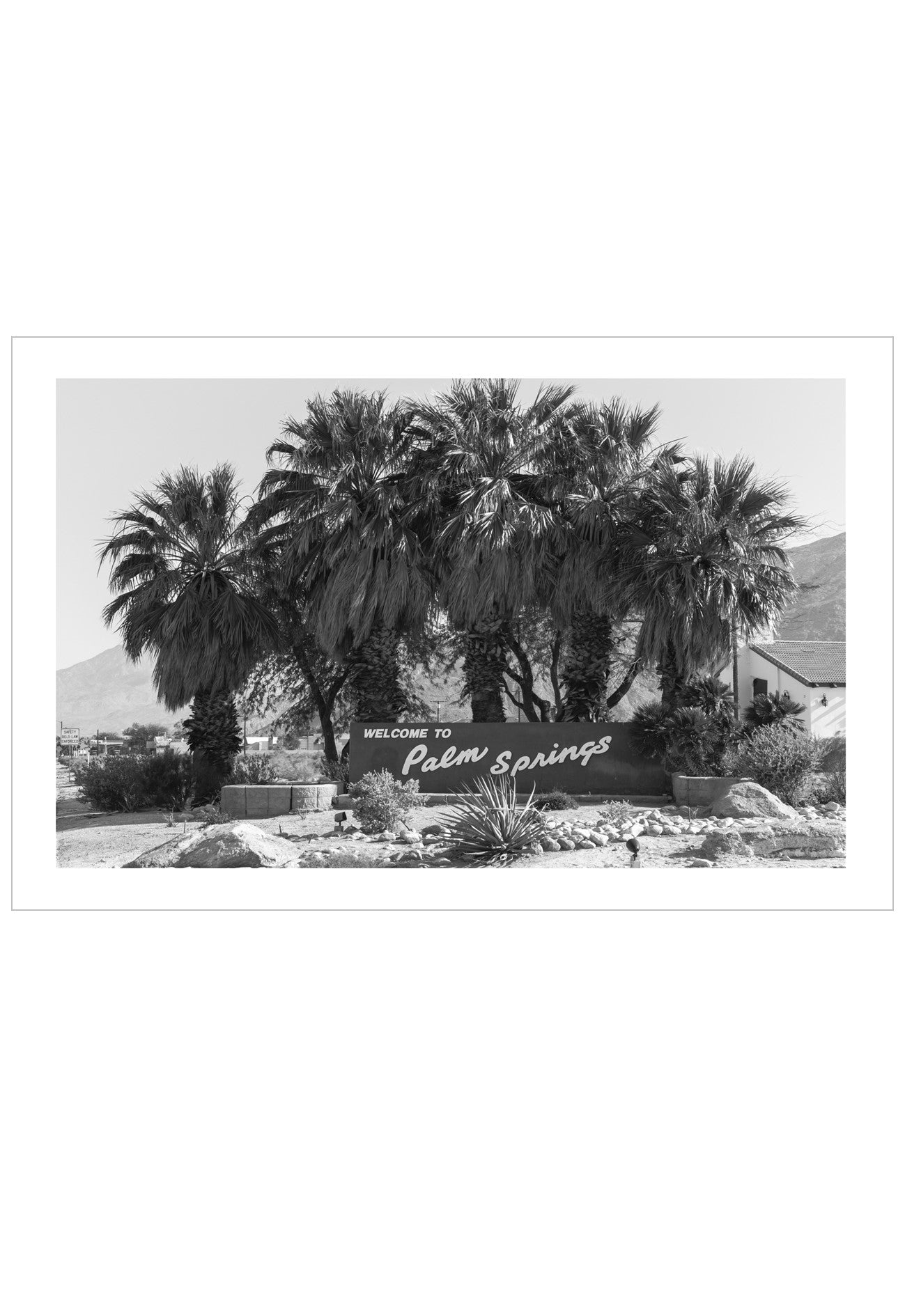 A black and white photographic art print of the 'Welcome to Palm Springs' sign, framed by tall palm trees, rocks, and desert mountains under a clear sky.