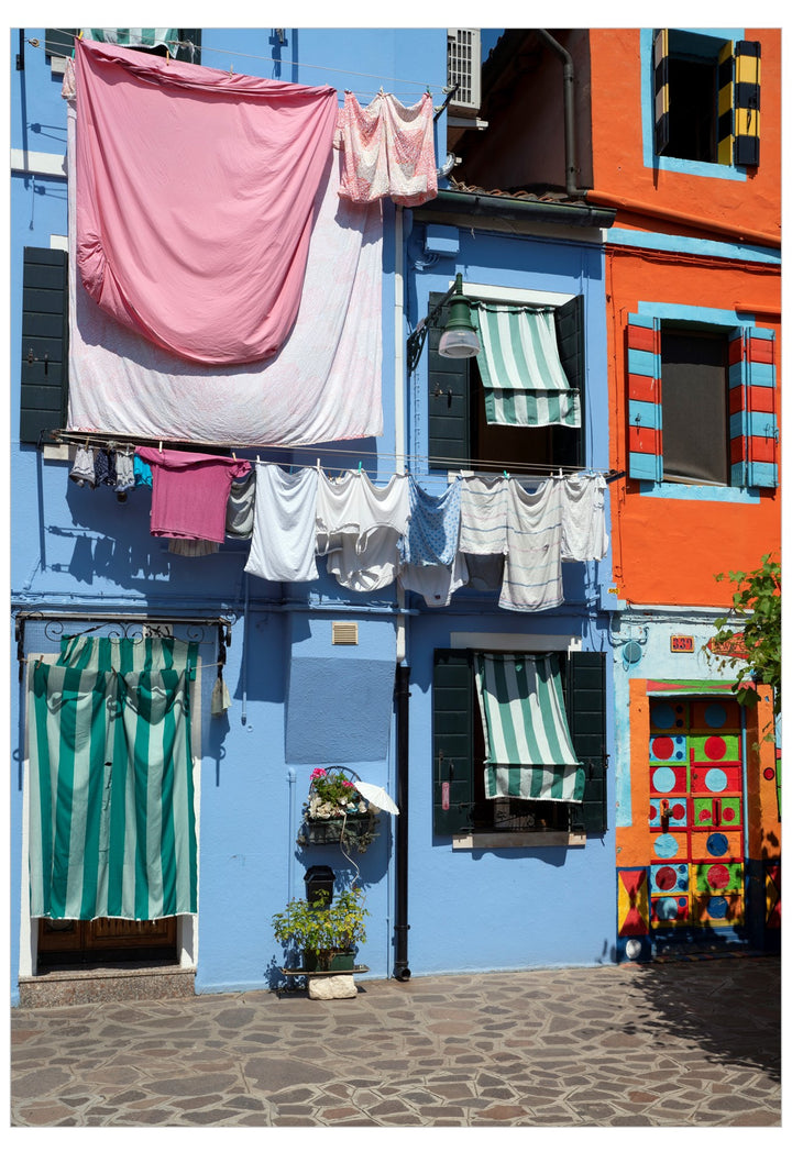 Colorful Italian village building facade with a large pink and white sheet hanging outside a blue house, alongside green striped awnings and an orange adjacent wall.