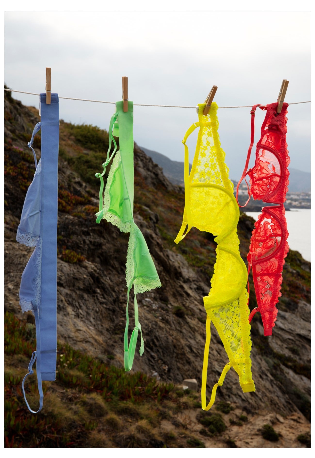 Four colorful lace lingerie sets in blue, green, yellow, and red hang from a clothesline with wooden clips against a rocky coastal cliff background.