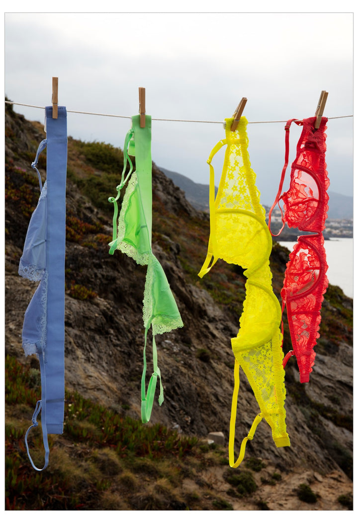 Four colorful lace lingerie sets in blue, green, yellow, and red hang from a clothesline with wooden clips against a rocky coastal cliff background.