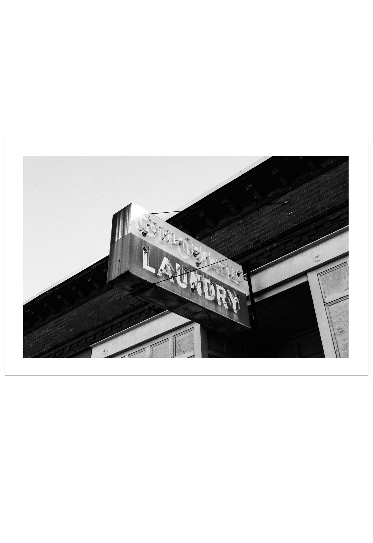 A black and white photographic print of a vintage wooden laundry sign hanging from a building eave, presented with a white border on a white background.