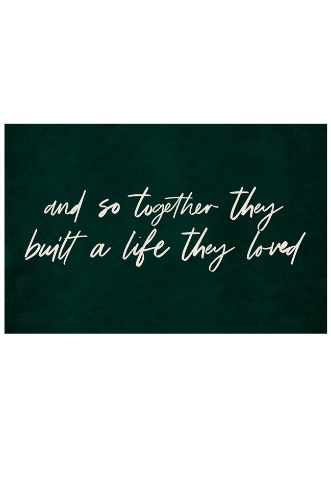 A rectangular dark green plaque or sign with white cursive text reading, 'and so together they built a life they loved,' centered on a white background.