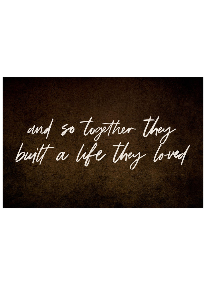 A rectangular wall-art style print with a dark brown textured background and white script text reading, 'and so together they built a life they loved.'.