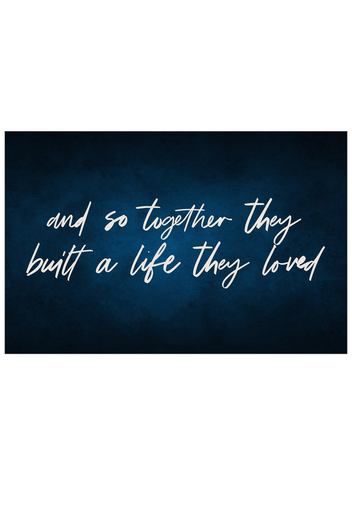 A rectangular wall-art style print with a dark navy textured background and white cursive text reading, 'and so together they built a life they loved.'.
