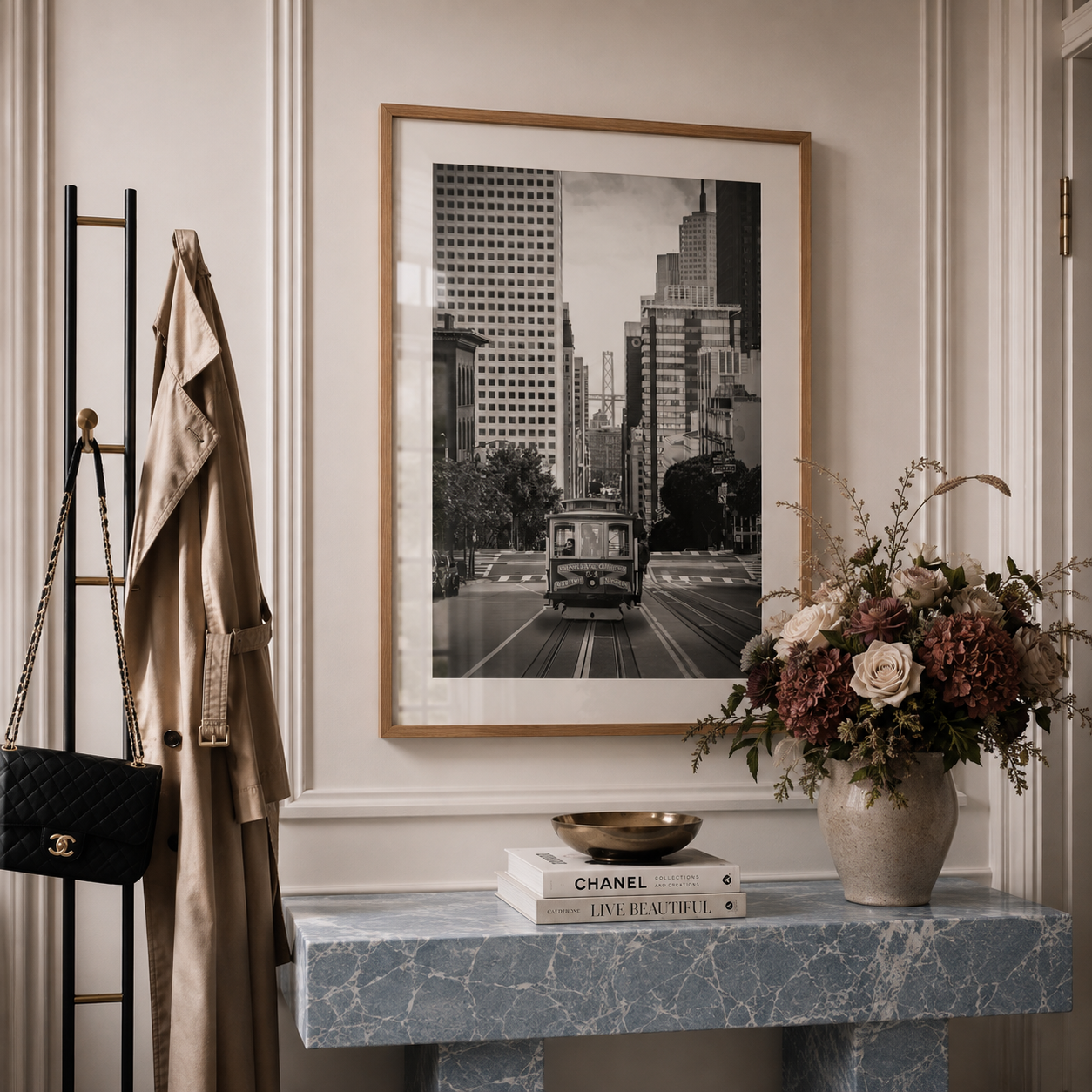 A framed black and white city street photograph with a vintage truck hangs on a cream wall above a blue marble console table. A beige trench coat, black quilted handbag, and a vase of dried flowers are arranged nearby in a styled interior setting.