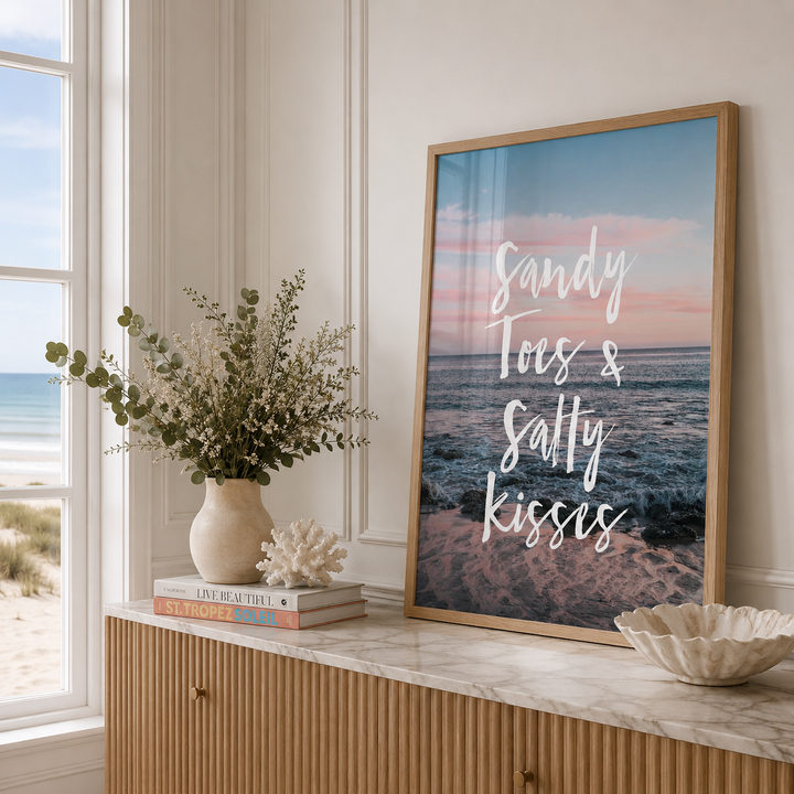 A framed beach-themed art print with the text 'Sandy Toes & Sally kisses' in white script over a sunset ocean scene, leaning on a marble-topped wooden cabinet in a bright room.