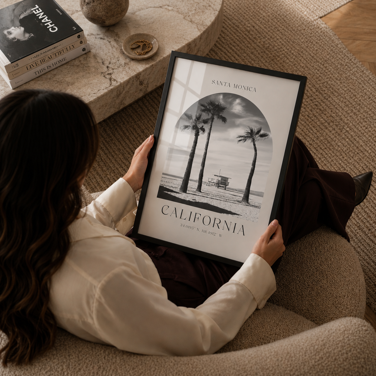 Person holding a framed print of palm trees and 'California' text in a cozy living room.