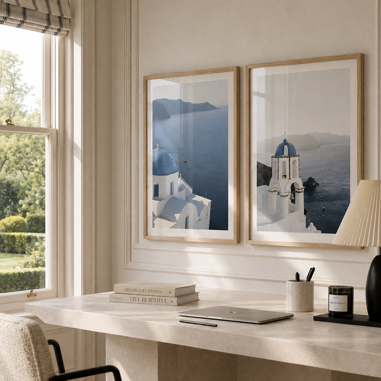 Two framed art prints depicting Santorini's white buildings and blue domes, displayed on a beige wall above a marble desk in a bright, modern room.