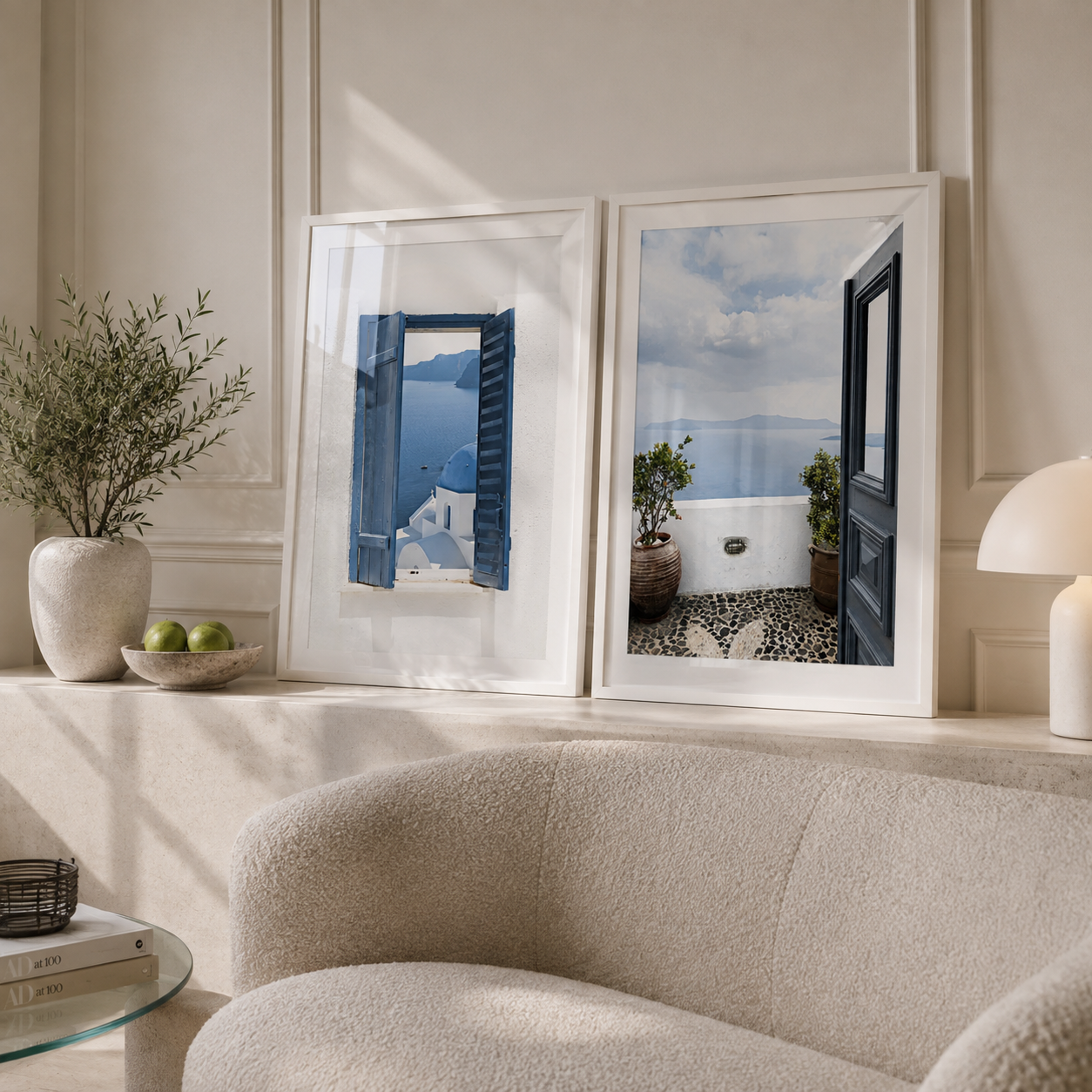 Two framed art prints depicting scenic Mediterranean views with blue shutters and a white wall, displayed on a beige mantelpiece in a styled living room setting.