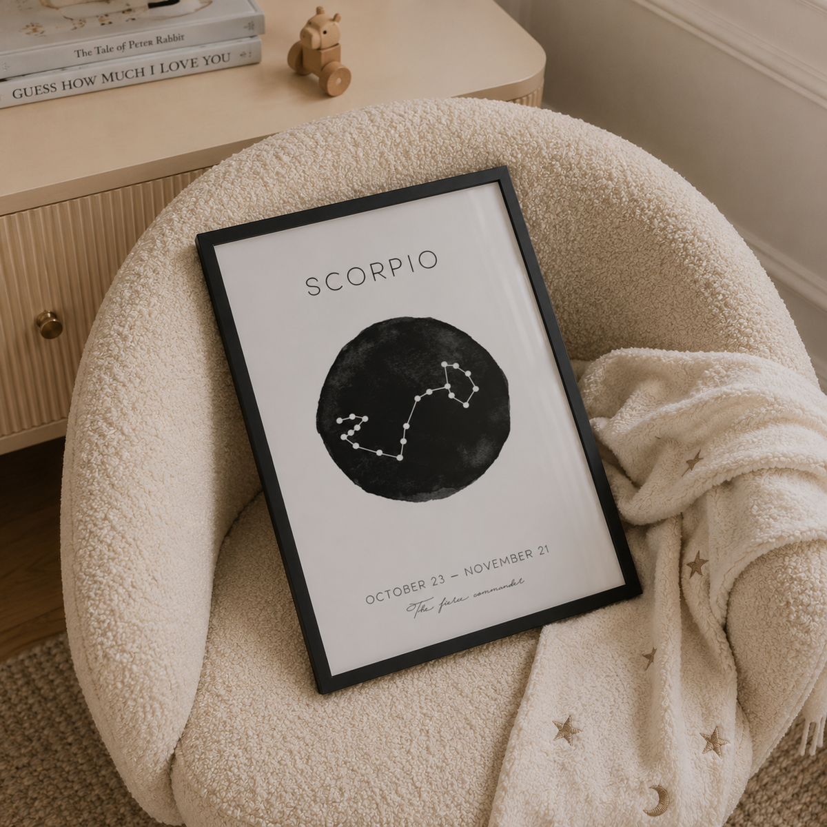 A framed Scorpio zodiac art print with a black circular astrological symbol on a white background, styled in a cozy cream-colored armchair with a knit blanket.