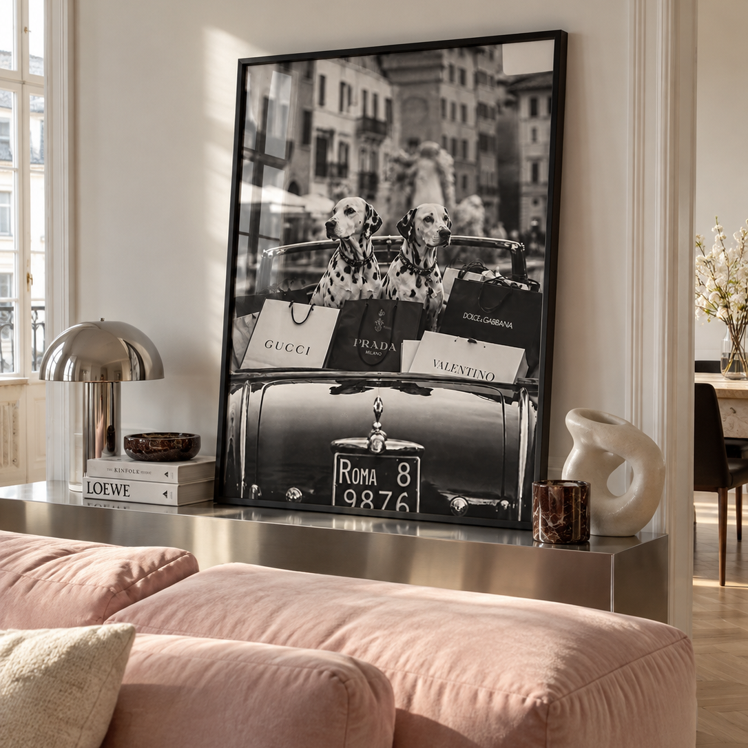 Black and white street photograph of two Dalmatian dogs sitting in the trunk of a vintage car, surrounded by luxury shopping bags from Gucci, Prada, Dolce & Gabbana, and Valentino, with a blurred city street and building facade in the background.