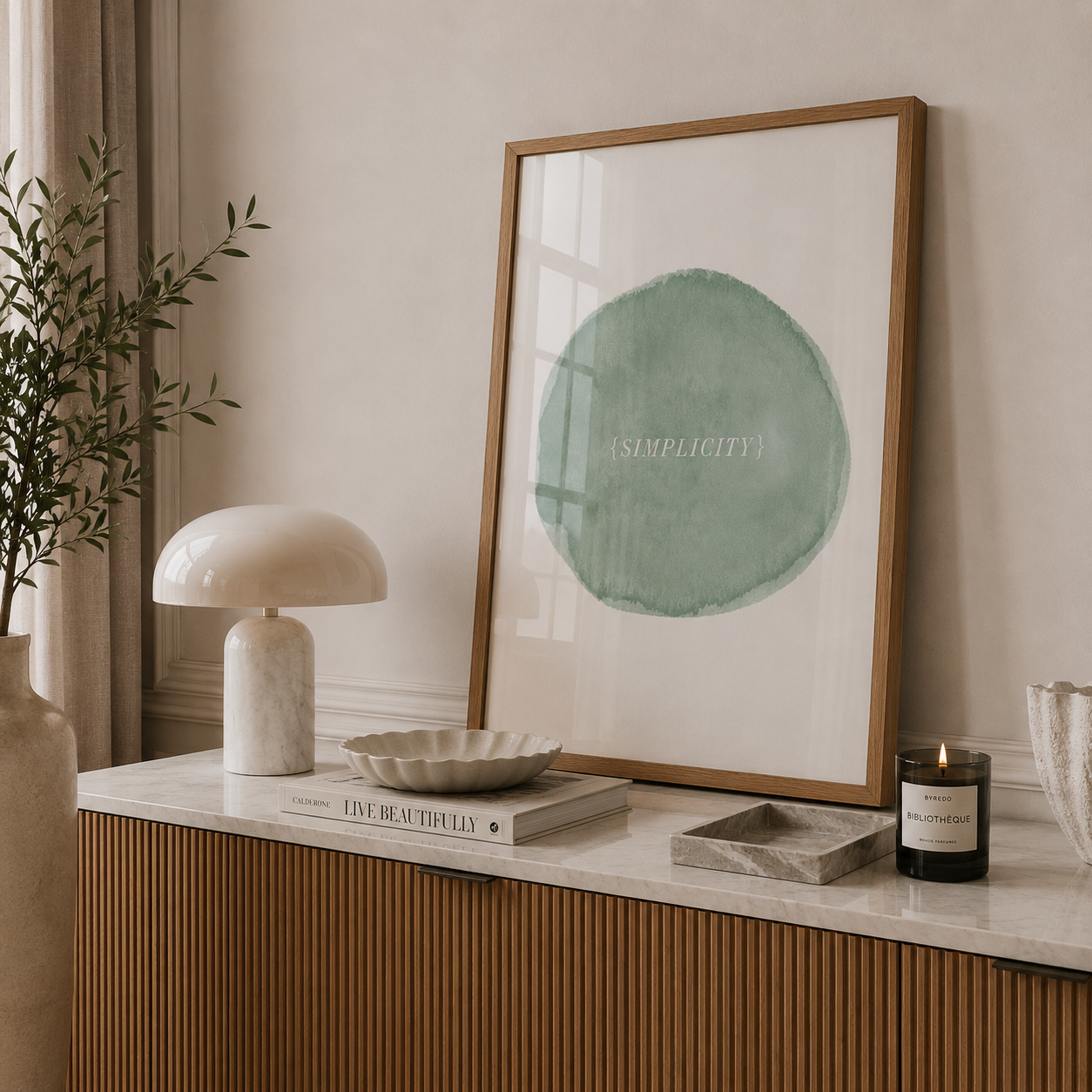 A framed minimalist art print featuring a sage green watercolor circle with the word 'SIMPLICITY' on a white background, styled in a warm, modern interior setting on a marble-topped console.