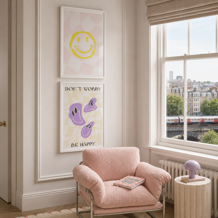 A styled interior room with a pink armchair, a small side table, and two framed wall art prints hanging above the chair. The lower print reads “DON’T WORRY BE HAPPY” with purple abstract shapes on a pale yellow background.