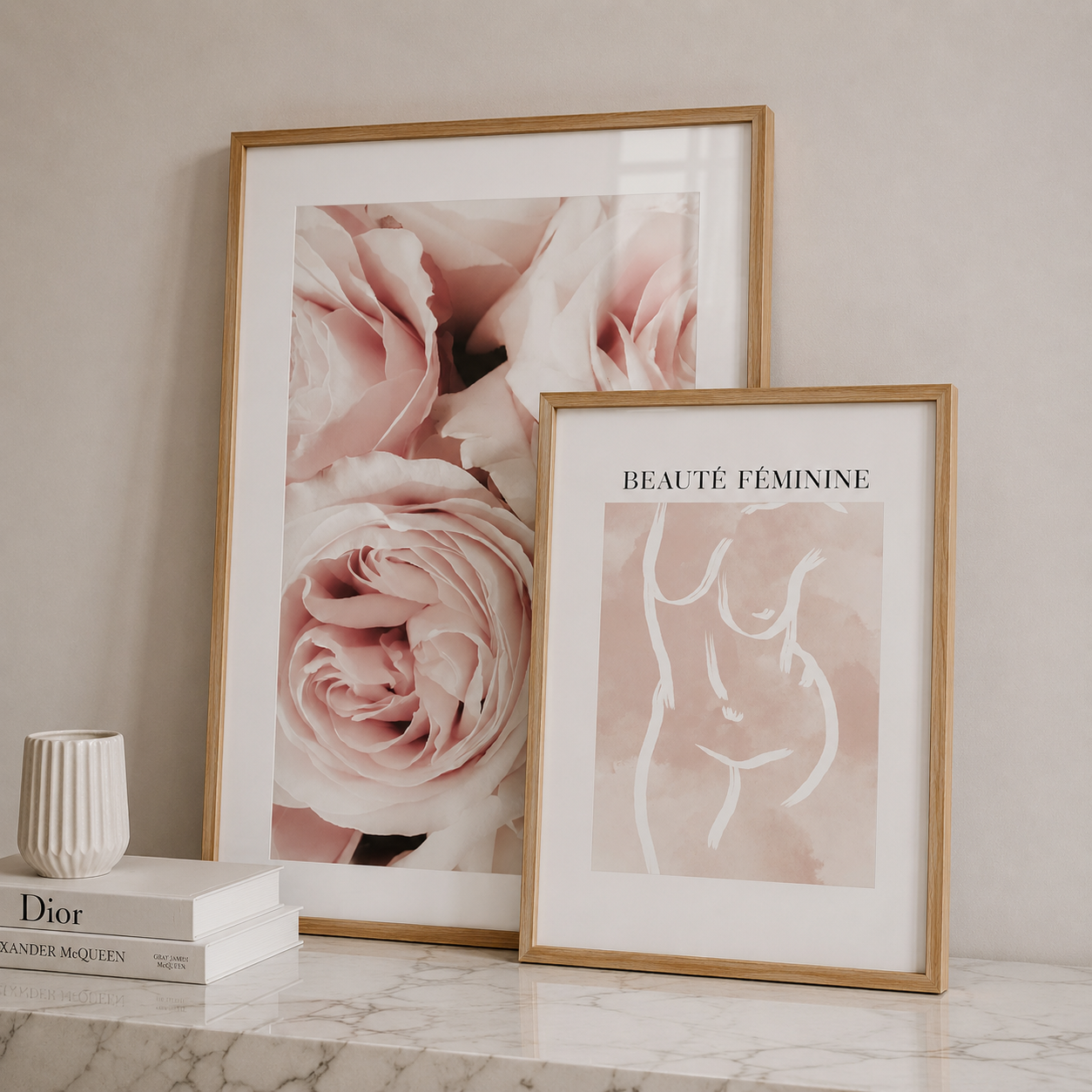 Two framed art prints displayed on a marble surface against a beige wall. The larger print features soft pink roses in a light wood frame, and the smaller print reads 'BEAUTÉ FEMININE' with a minimalist white line drawing of a female torso on a blush pink background in a matching frame.