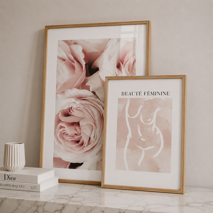 Two framed art prints displayed on a marble surface against a beige wall. The larger print features soft pink roses in a light wood frame, and the smaller print reads 'BEAUTÉ FEMININE' with a minimalist white line drawing of a female torso on a blush pink background in a matching frame.