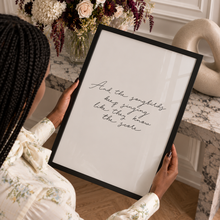 A person holds a black-framed art print featuring elegant black cursive script on a white background, set against a warm, styled interior with a floral arrangement and marble surfaces.