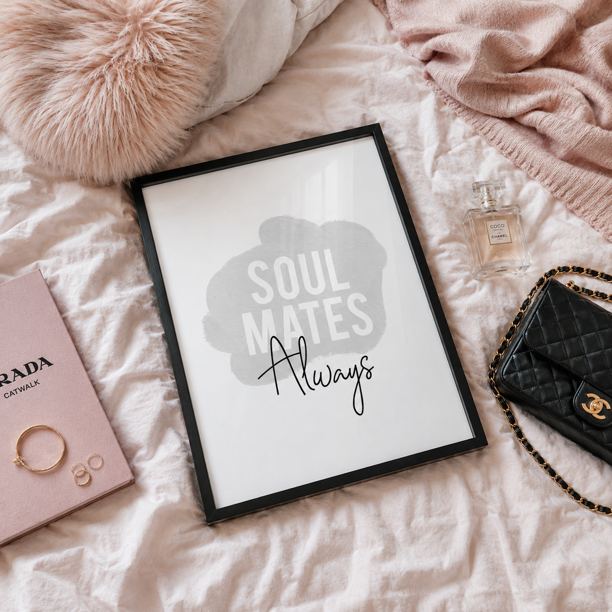 A framed wall art print reading 'SOUL MATES Always' in gray and black lettering on a white background, styled on a pink bed with a fluffy pillow, pink blanket, perfume bottle, jewelry, and a black quilted handbag nearby.