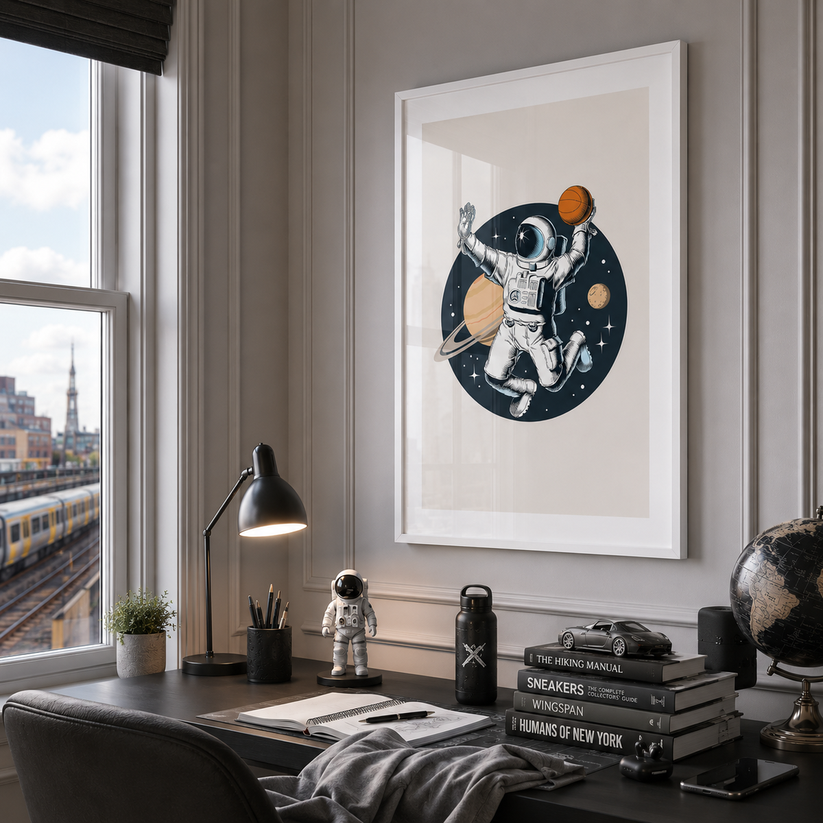 A framed art print featuring an astronaut floating in space with planets, displayed on a gray paneled wall above a modern desk setup with a lamp, books, and a globe.