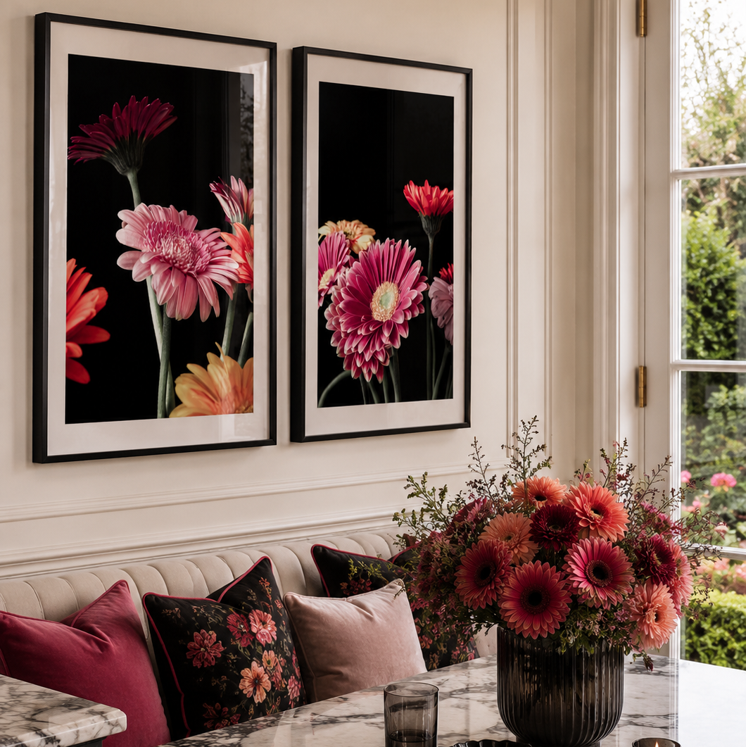 A styled interior scene with two large framed floral wall art prints above a tufted sofa. The prints feature bold pink, red, orange, and yellow flowers on black backgrounds with white mats and black frames.