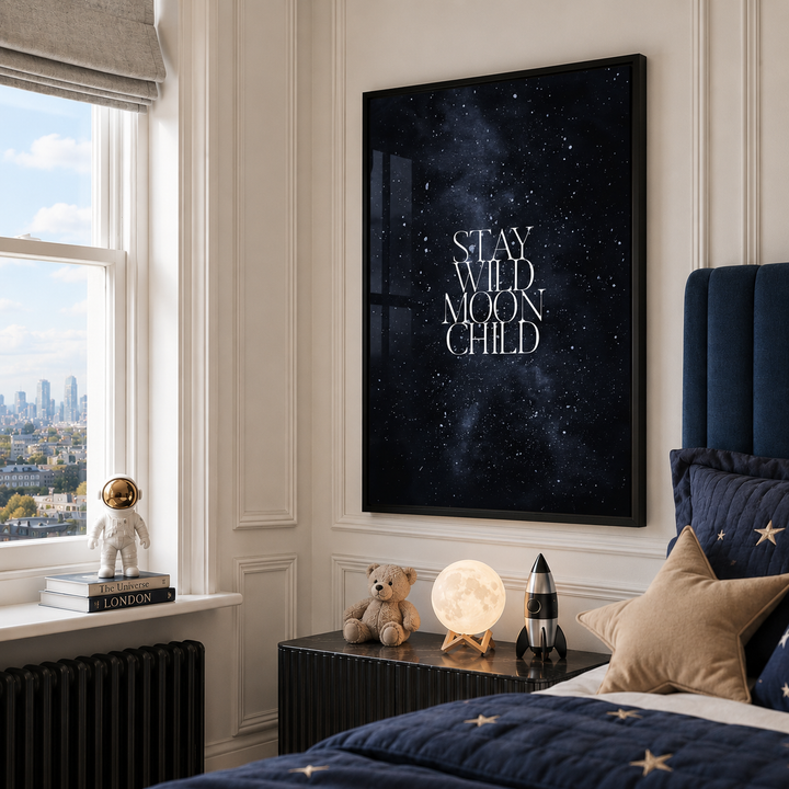 A framed vertical wall art print with the phrase “STAY WILD MOON CHILD” in white serif text over a dark starry night sky, displayed in a styled bedroom above a bedside table.