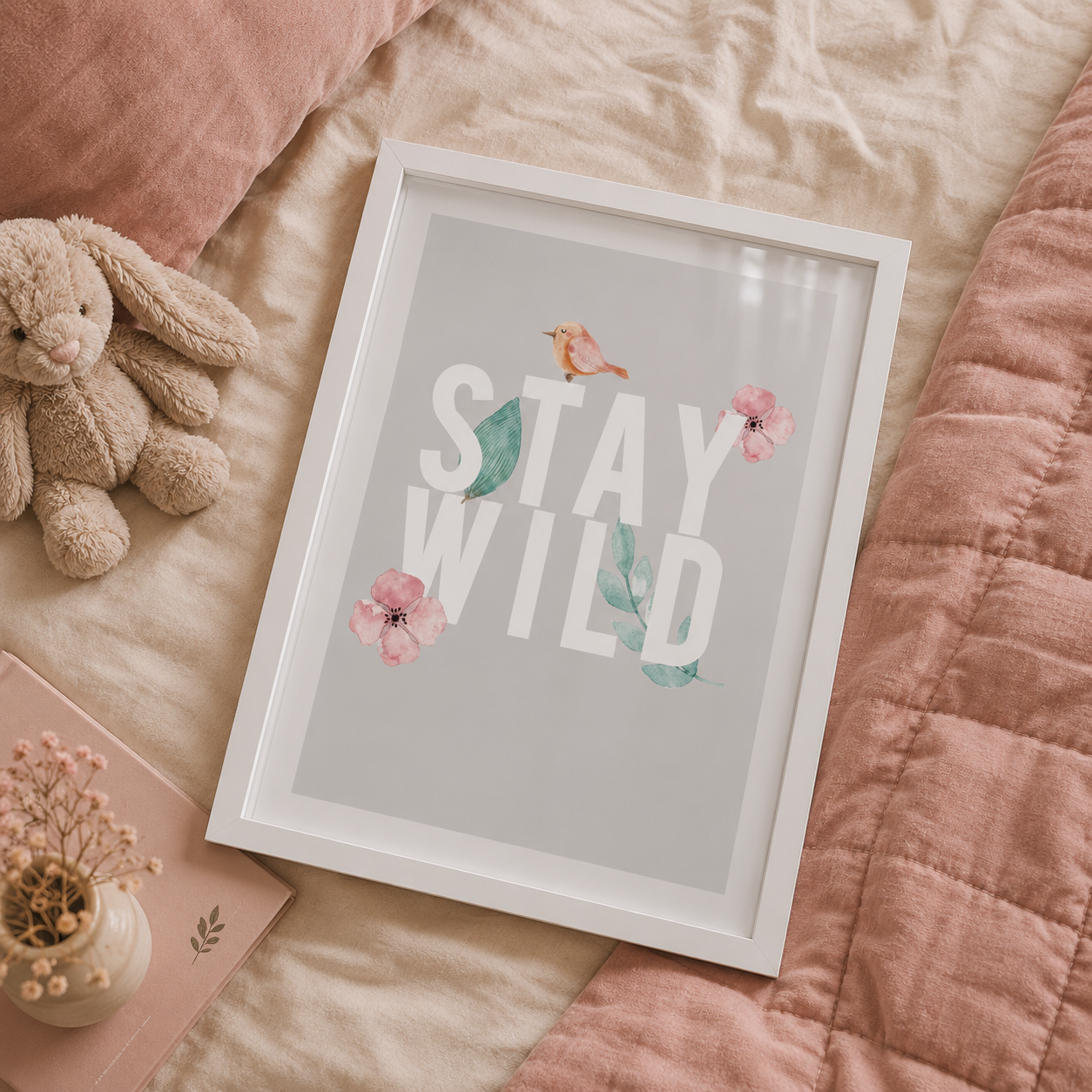 A framed wall art print reading “STAY WILD” in large white letters, decorated with pink flowers, teal leaves, and a small bird, styled on a bed with a plush bunny, pillow, and quilted blanket.