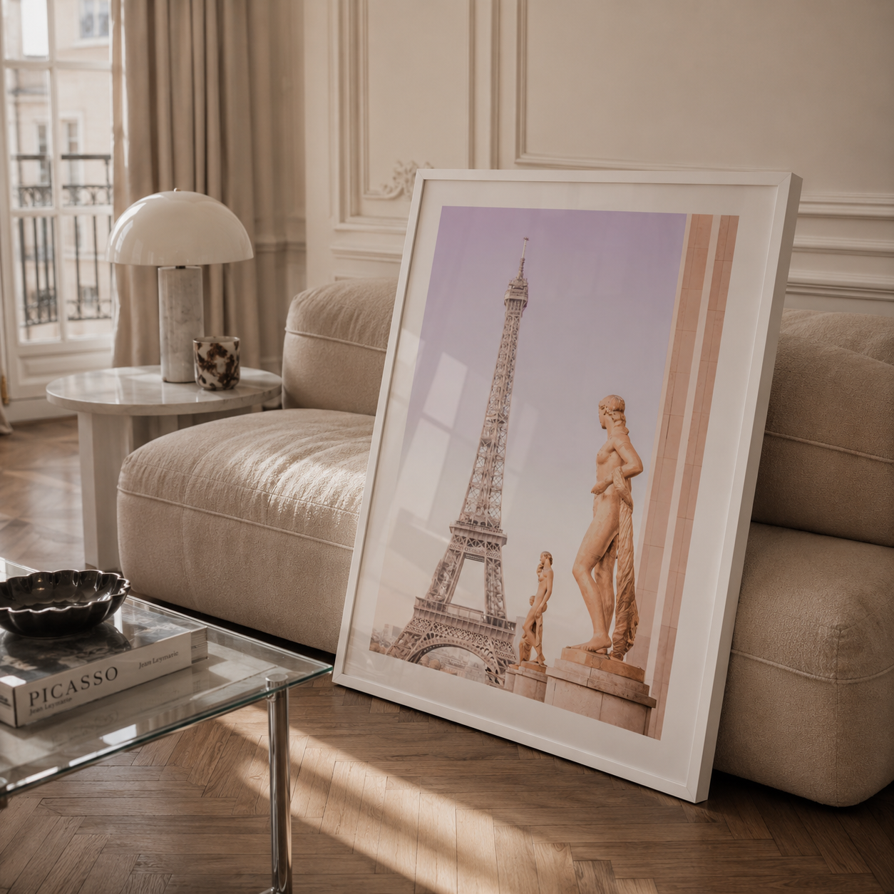 A framed art print leaning against a beige sofa in a stylishly decorated living room. The print features a pastel-colored photograph of the Eiffel Tower and a classical statue, enclosed in a clean white frame.
