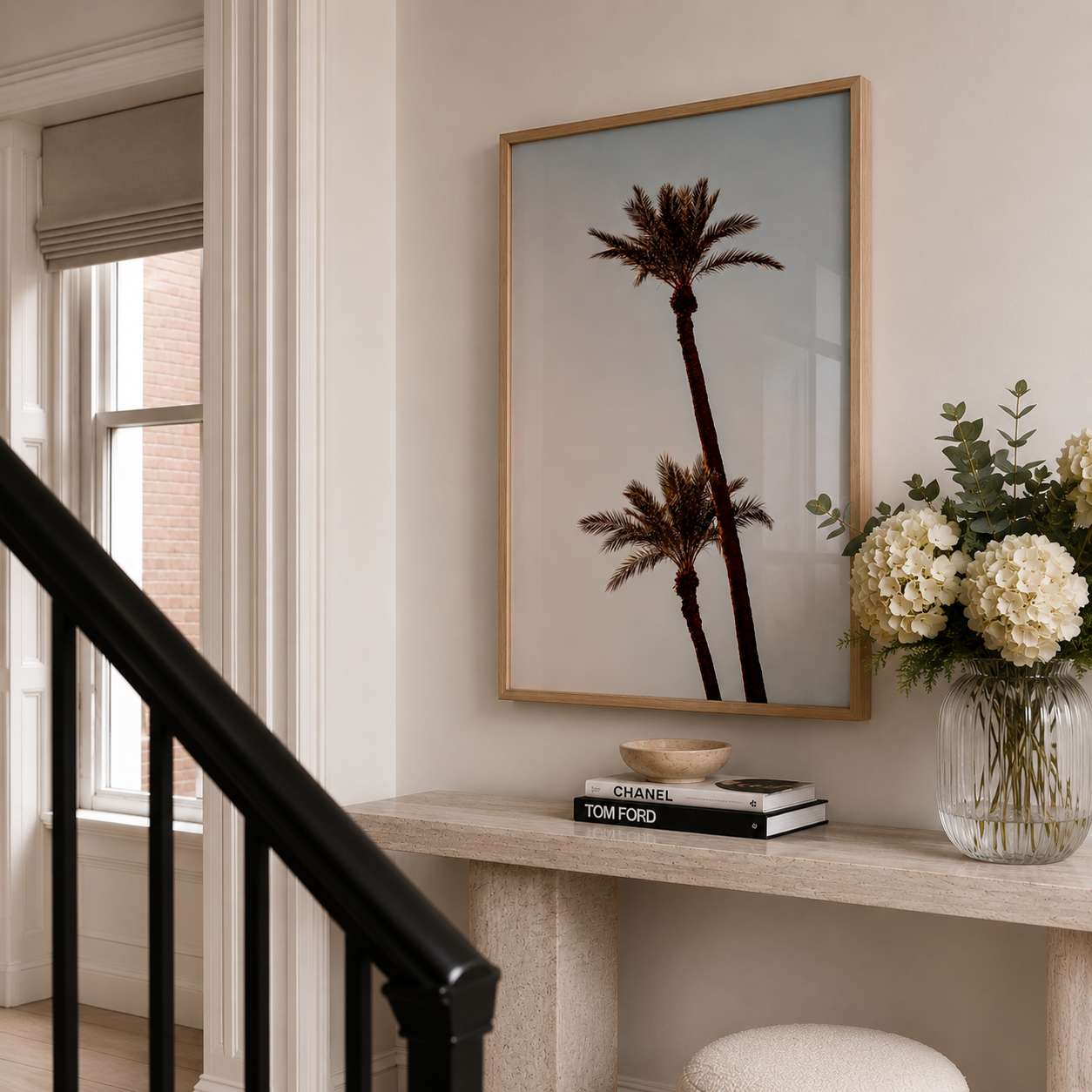 A framed art print featuring two palm trees hanging on a beige wall above a console table with books and a vase of flowers. The print is enclosed in a thin gold frame.