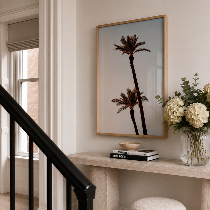A framed art print featuring two palm trees hanging on a beige wall above a console table with books and a vase of flowers. The print is enclosed in a thin gold frame.