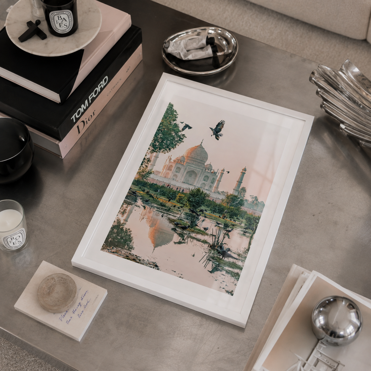 A framed art print depicting the Taj Mahal with a bird silhouette and soft pink watercolor wash, styled on a gray tabletop with books, candles, and metallic accessories.