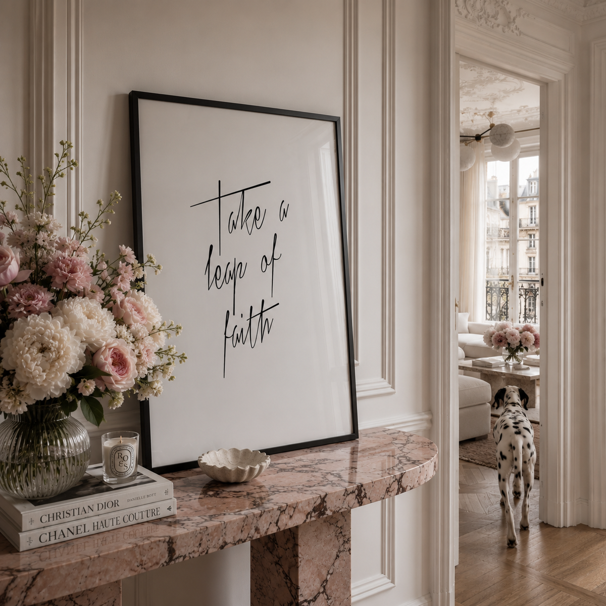 A framed wall art print with the phrase 'Take a leap of faith' in black script on a white background, styled on a pink marble console table in a elegant interior. A vase of pink and white flowers, a candle, a small dish, and a Dalmatian dog are visible in the room.