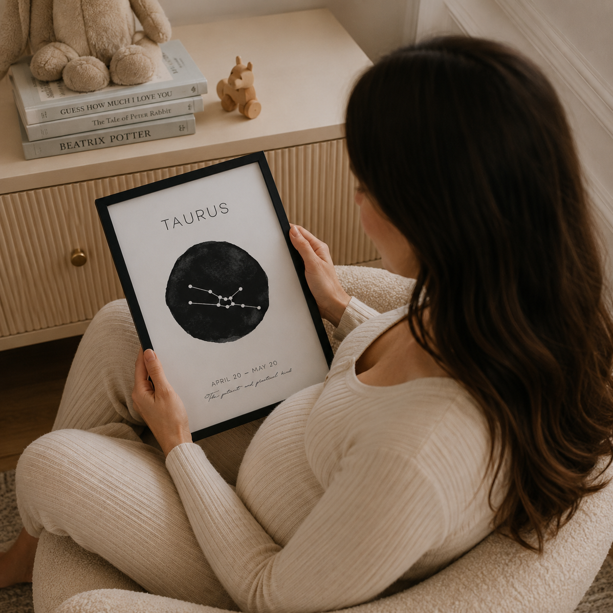 A woman in a cozy cream-colored ribbed sweater and matching pants sits in a plush armchair, holding a framed Taurus zodiac sign print. The print features a black circle with white constellation lines and text.