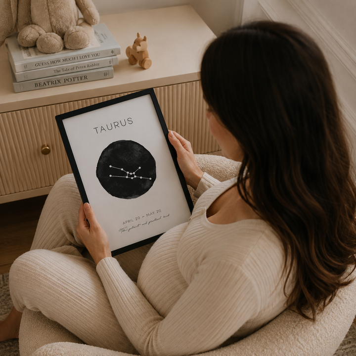 A woman in a cozy cream-colored ribbed sweater and matching pants sits in a plush armchair, holding a framed Taurus zodiac sign print. The print features a black circle with white constellation lines and text.