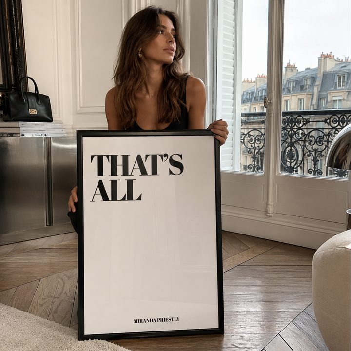 A woman holds a large framed art print with the text 'THAT'S ALL' in bold black letters on a white background. 