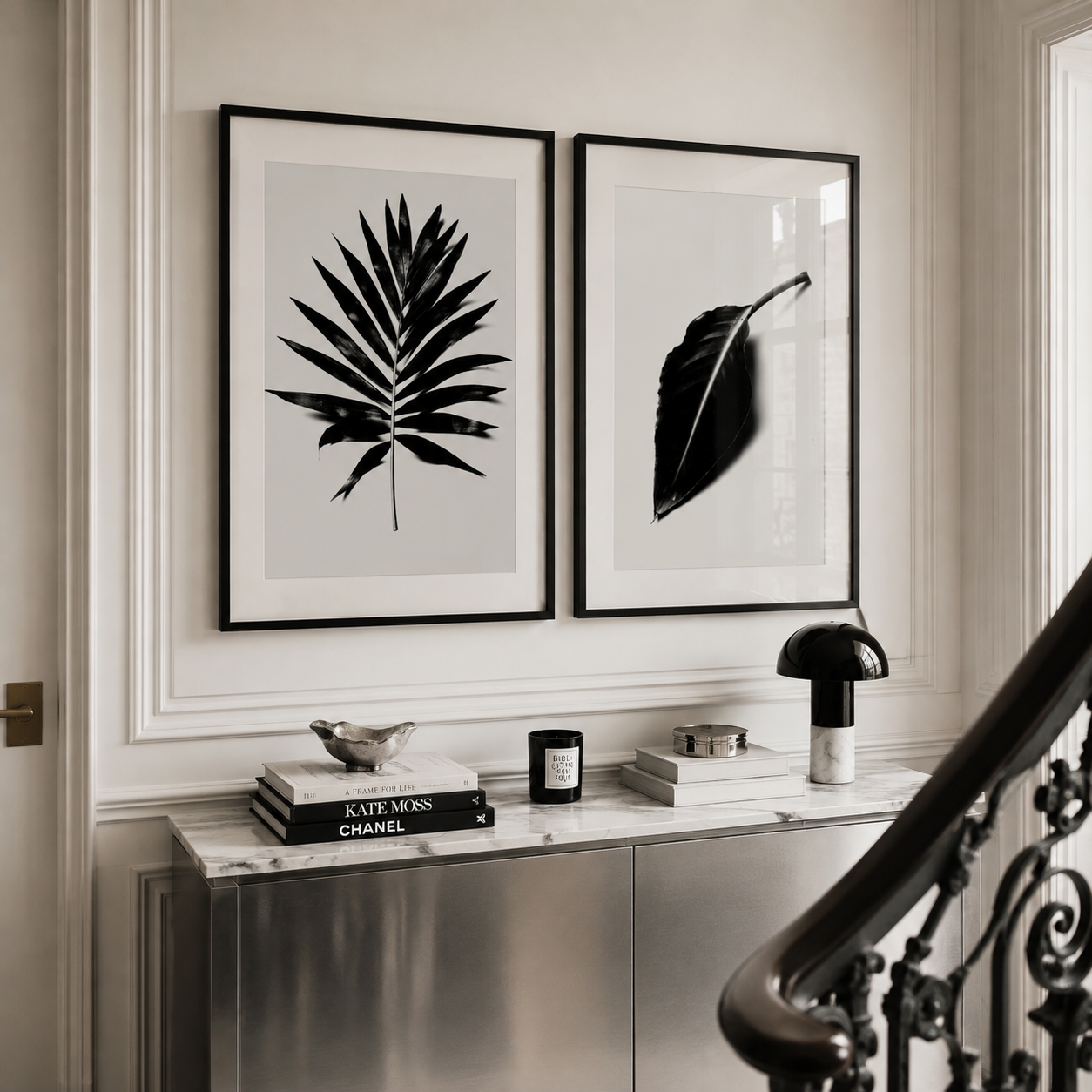 Two framed black and white botanical art prints hanging side-by-side on a white paneled wall above a marble console table with decorative items and a staircase railing in the foreground.