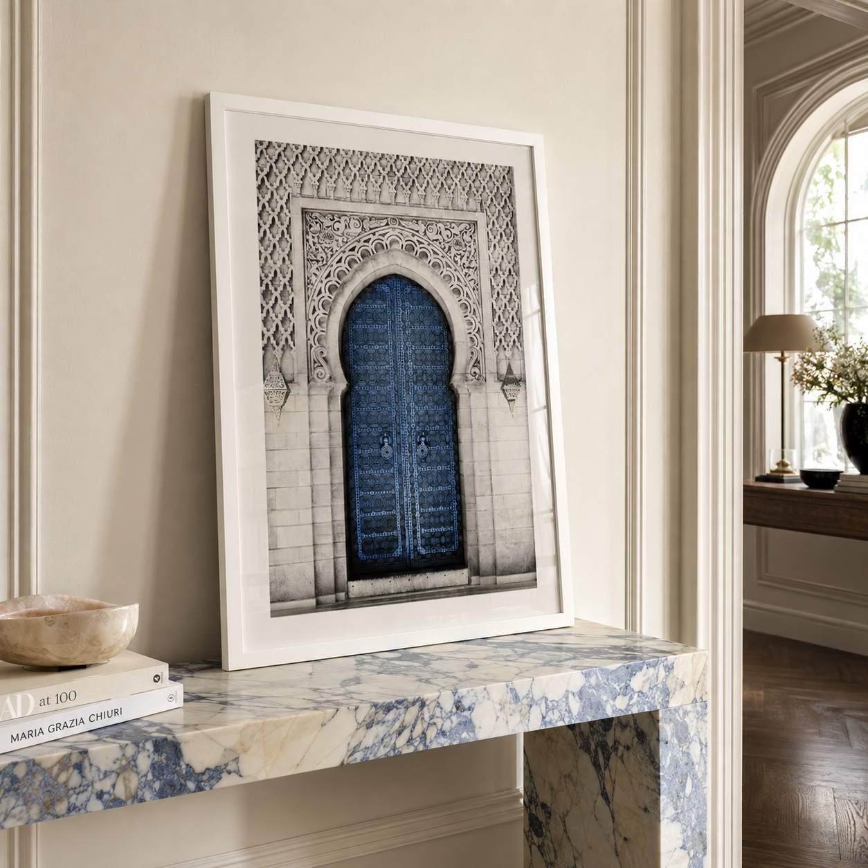 A framed art print featuring a blue Moroccan-style wooden door set within an ornate gray stone archway, displayed leaning against a beige wall on a blue and white marble console table.