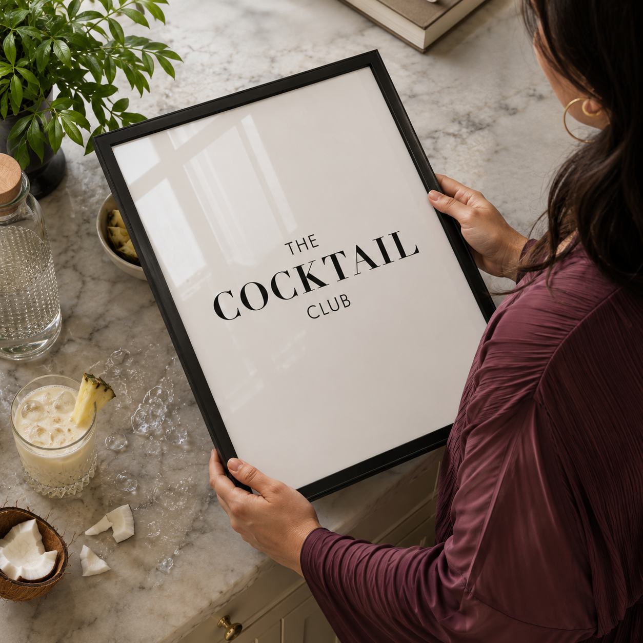 Person holding a framed poster titled 'The Cocktail Club' in a kitchen setting with a drink and plant.
