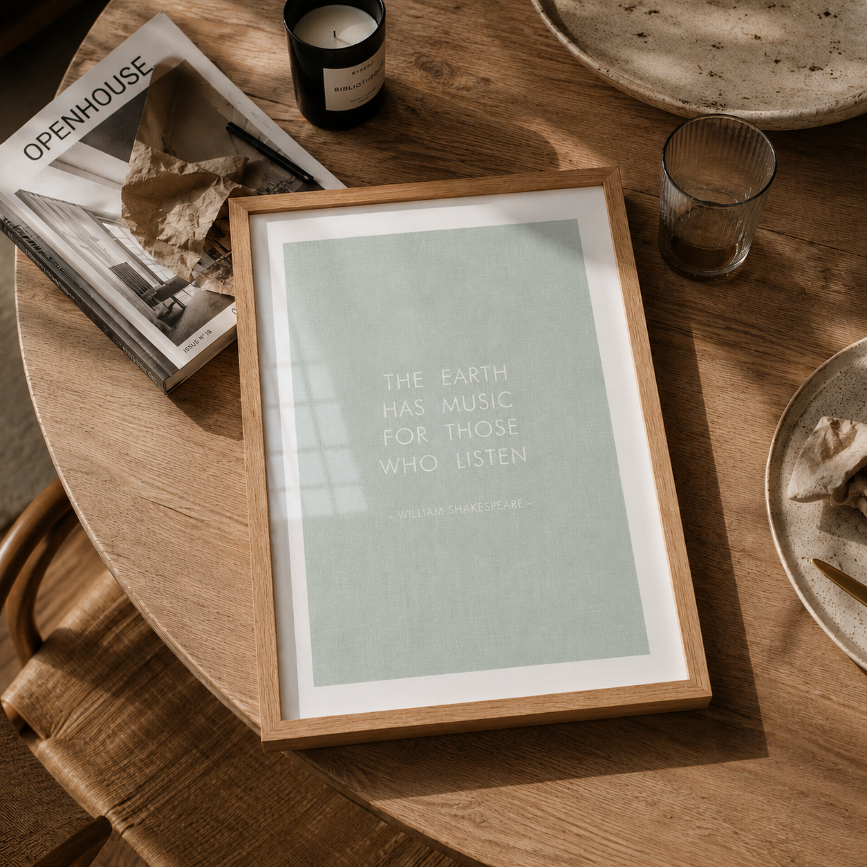 A framed art print featuring a quote by William Shakespeare, displayed on a rustic wooden table alongside a candle, magazine, and other home decor items.