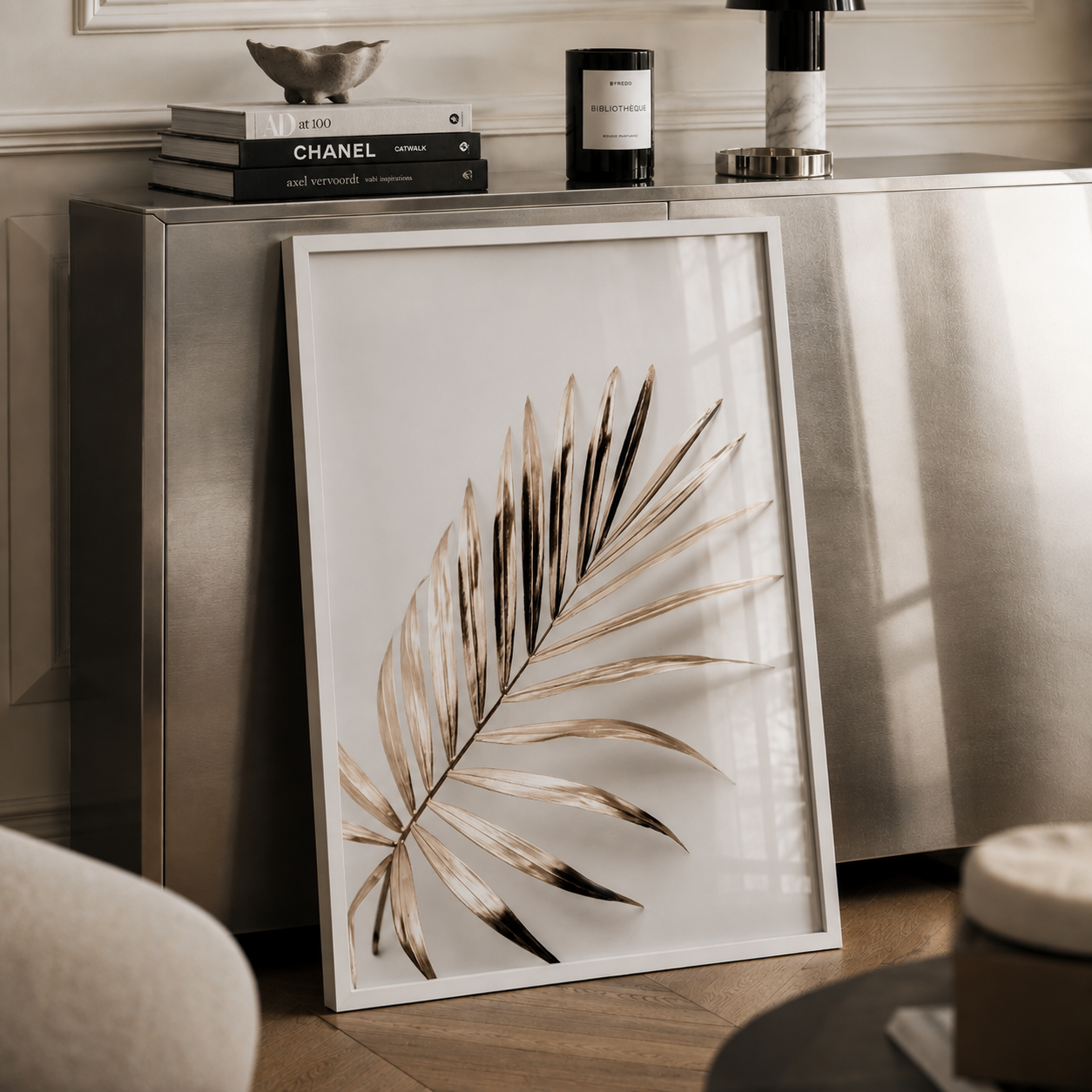 A framed botanical art print featuring a dried palm leaf in warm beige and brown tones, leaning against a metallic console table in a styled interior setting.