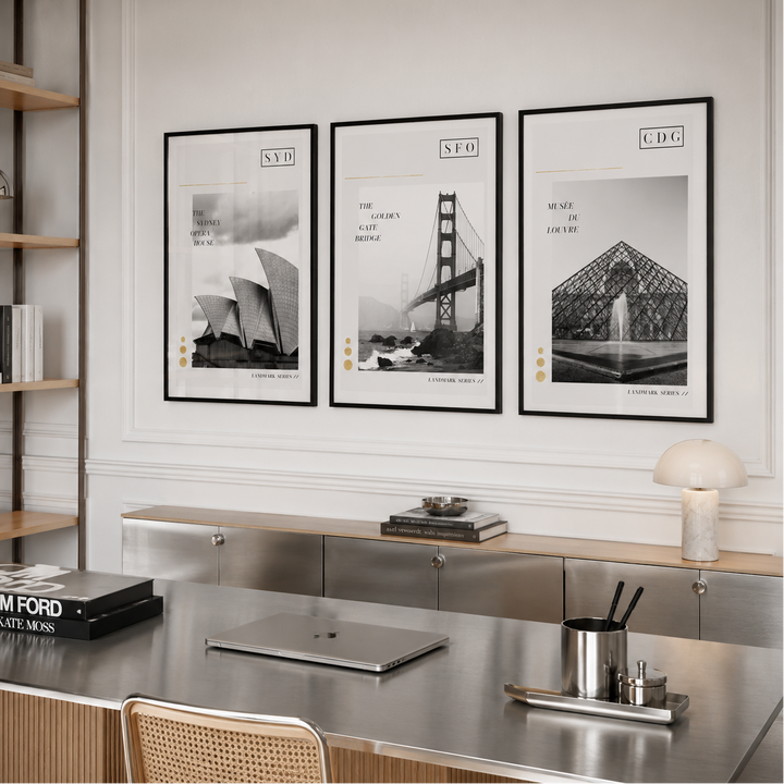 Three framed black-and-white architectural art prints hang on a white wall above a modern desk in a styled interior room. The prints feature the Sydney Opera House, Golden Gate Bridge, and Louvre Pyramid with stylized city labels.