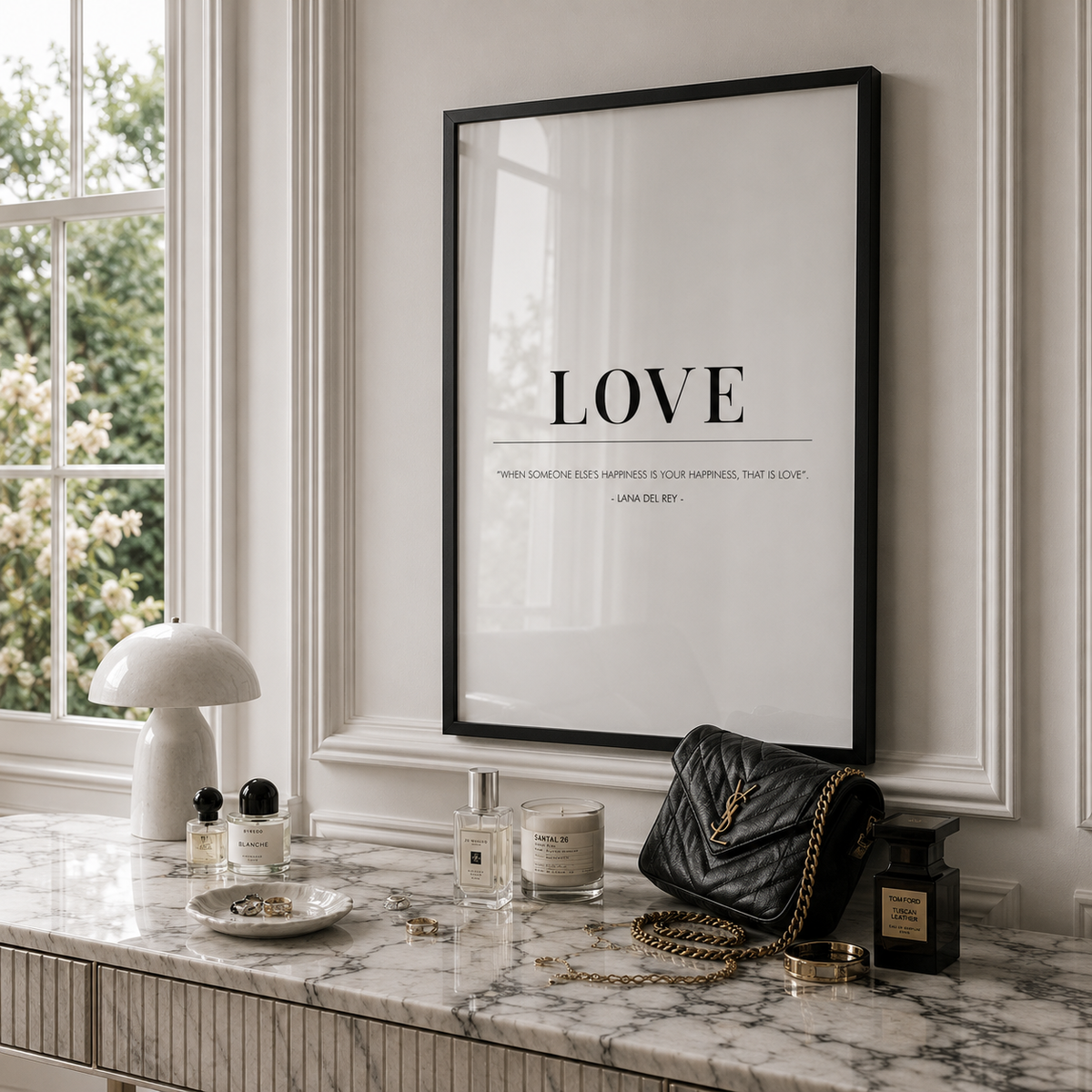 A framed wall art print with the word LOVE and a quote in black text on a white background, hanging on a light gray paneled wall above a marble console table styled with a lamp, candle, perfume, and a black quilted handbag.