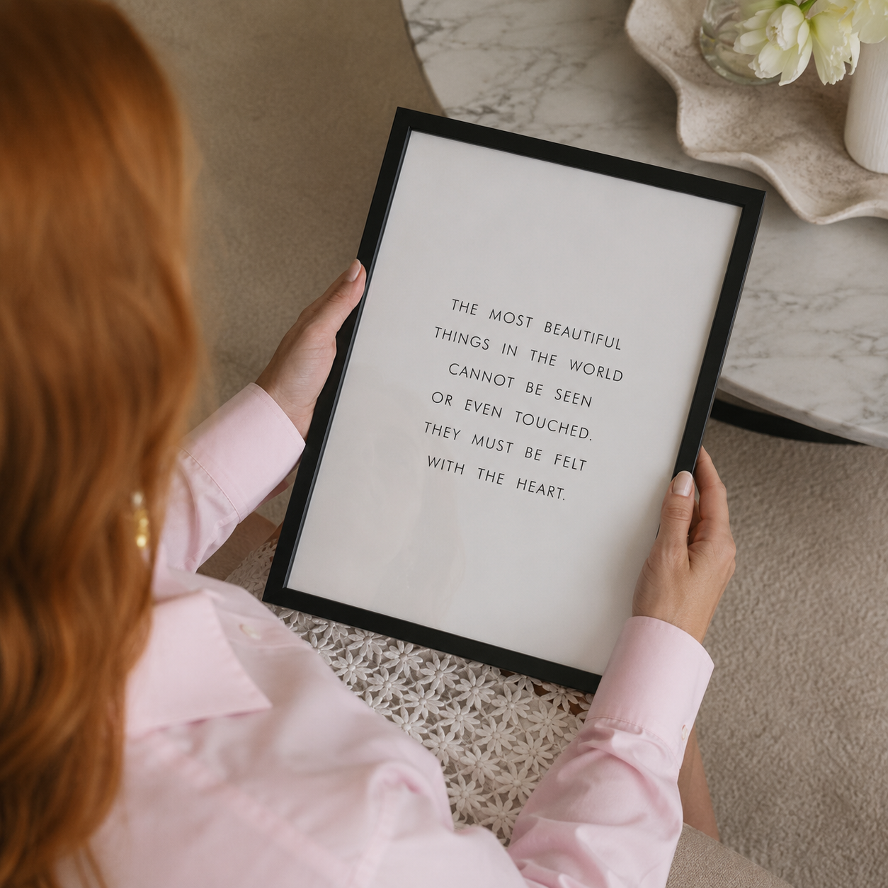 A person in a light pink Dior shirt holds a framed art print with a black frame. The print features a quote in black text on a white background.