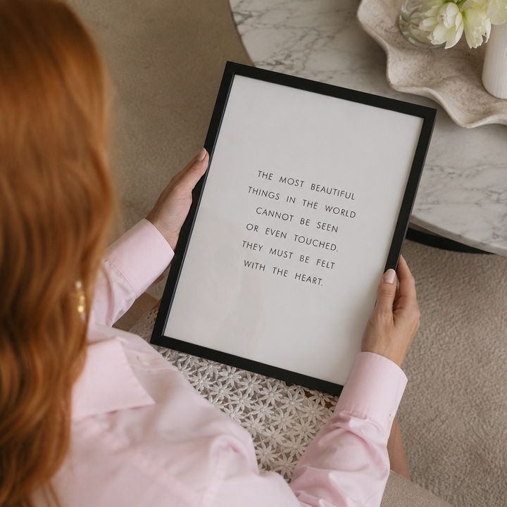 A person in a light pink Dior shirt holds a framed art print with a black frame. The print features a quote in black text on a white background.