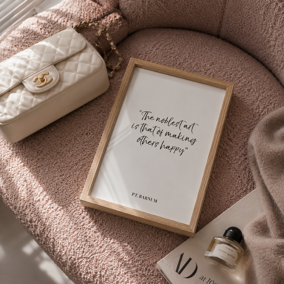 A framed art print with the quote 'The noblest art is that of making others happy' by Pt. Raimum, styled on a dusty pink textured chair next to a cream quilted handbag and a perfume bottle.